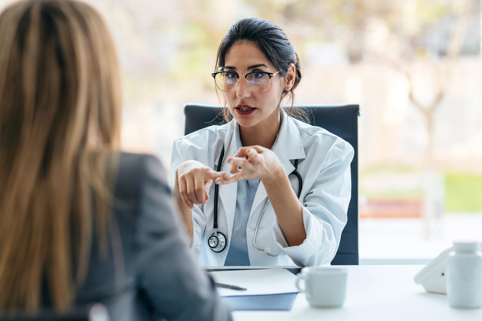 Clinician with glasses and a stethoscope speaking emphatically with a patient across a desk