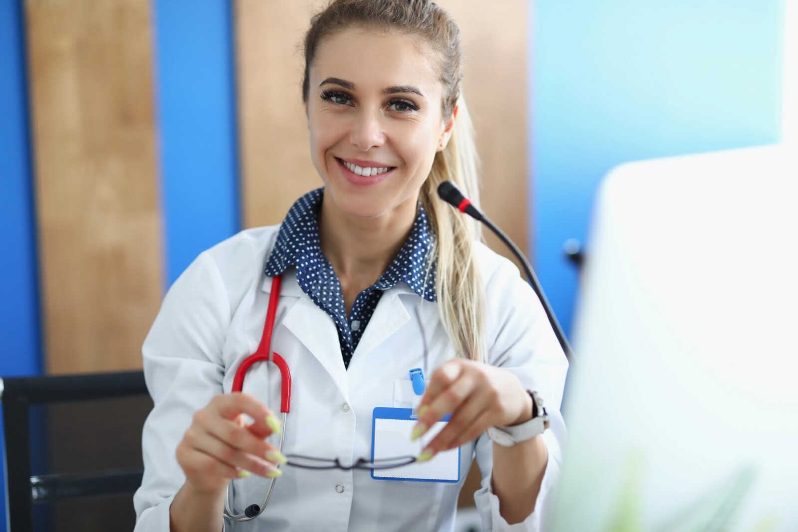 Smiling clinician with a red stethoscope holding her glasses at a desk with a microphone and computer