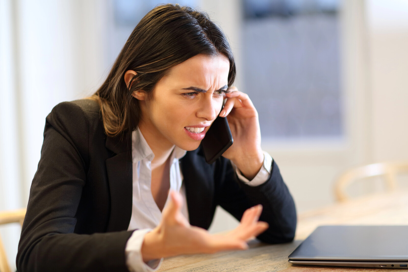 Frustrated woman gesturing while arguing on a phone call at a table with a laptop