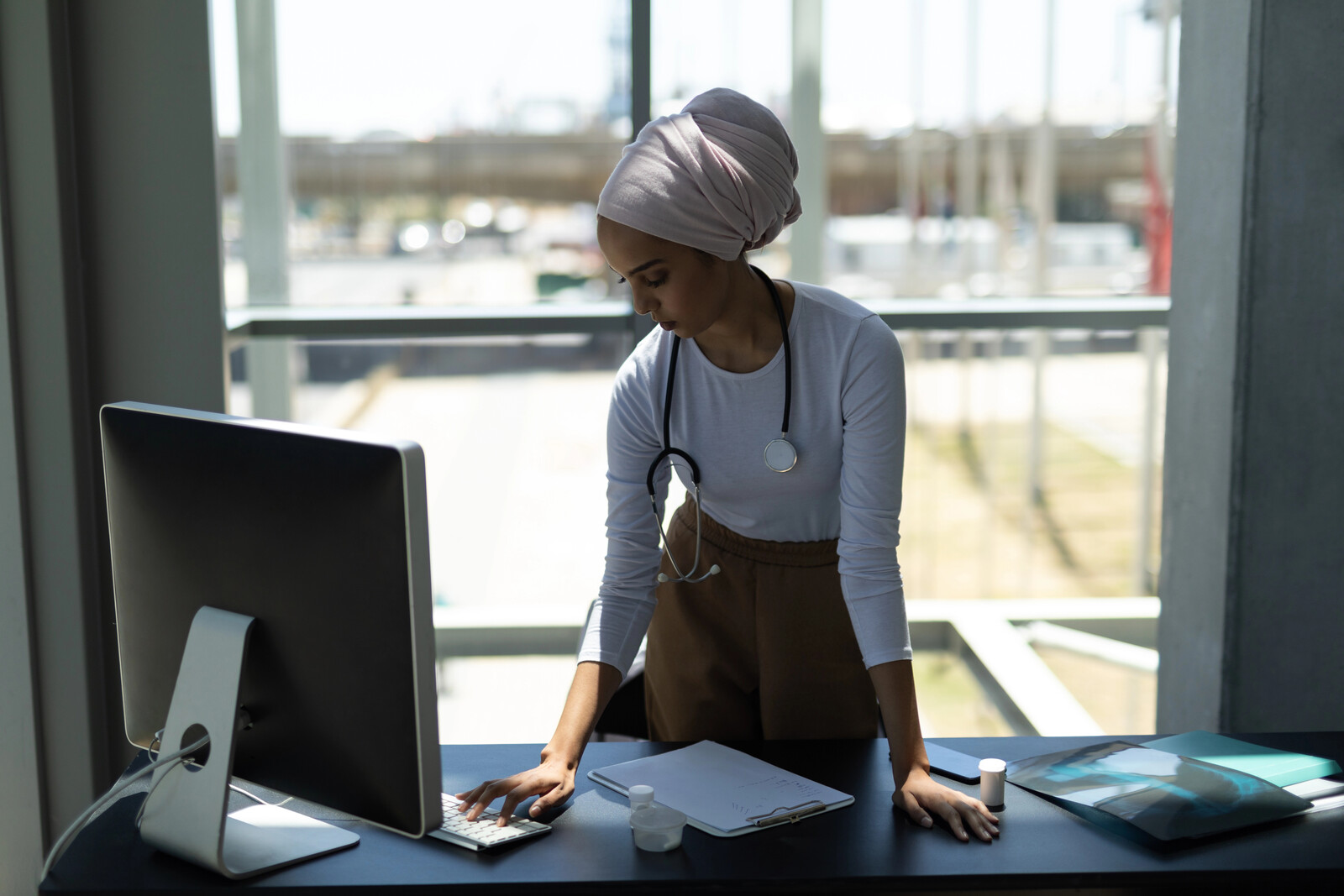 Clinician in a headwrap with a stethoscope standing at a desk, typing on a keyboard beside paperwork