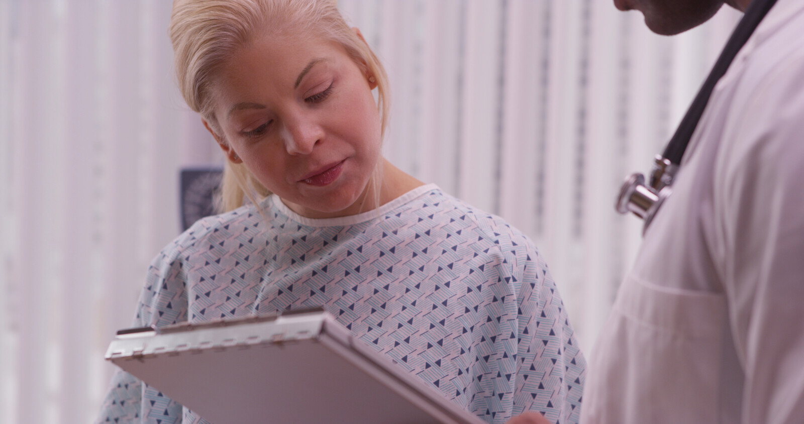 Patient in a hospital gown reading notes on a clipboard held out by a clinician