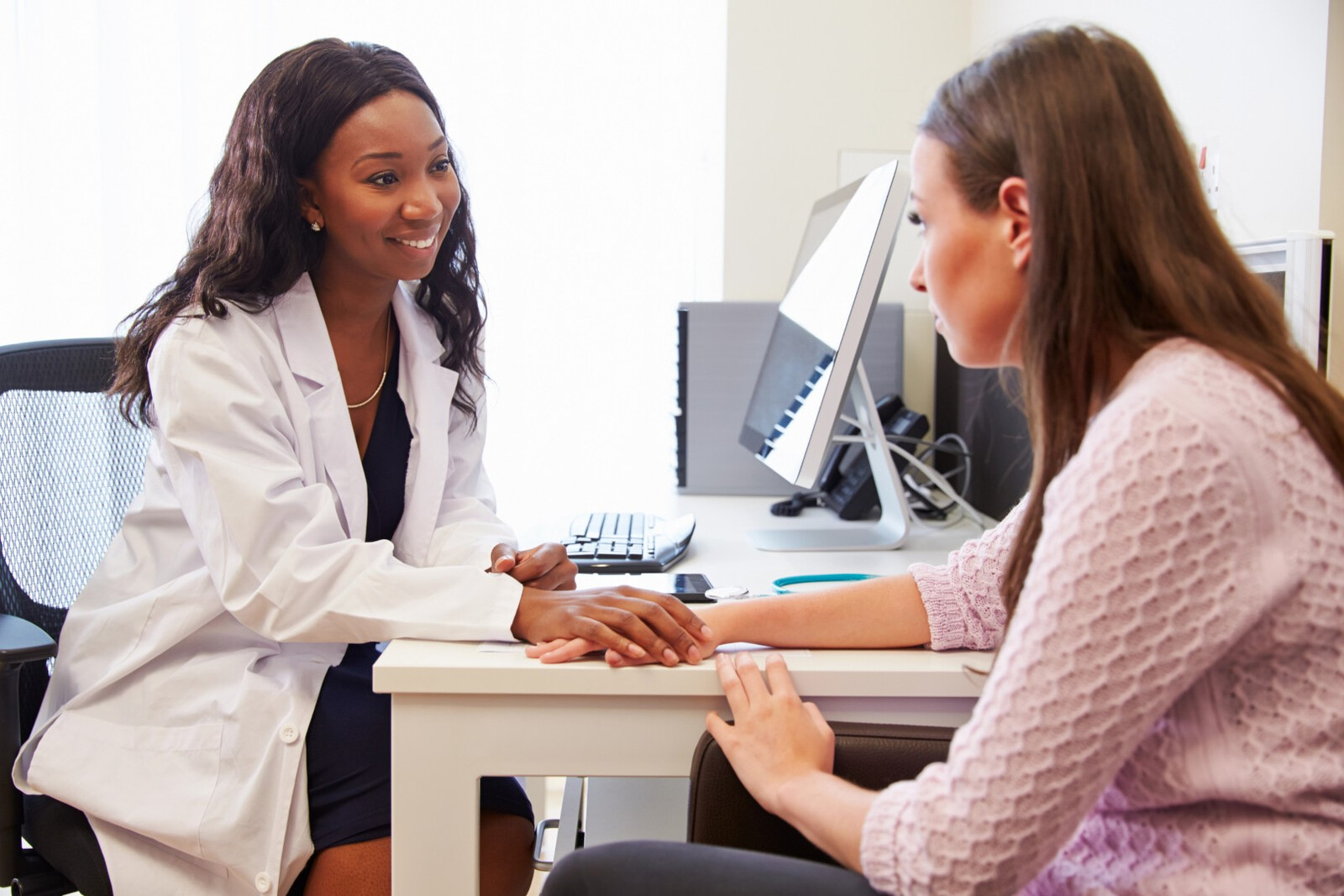 A clinician in a white coat sits across a desk from a patient, smiling and resting a reassuring hand on hers
