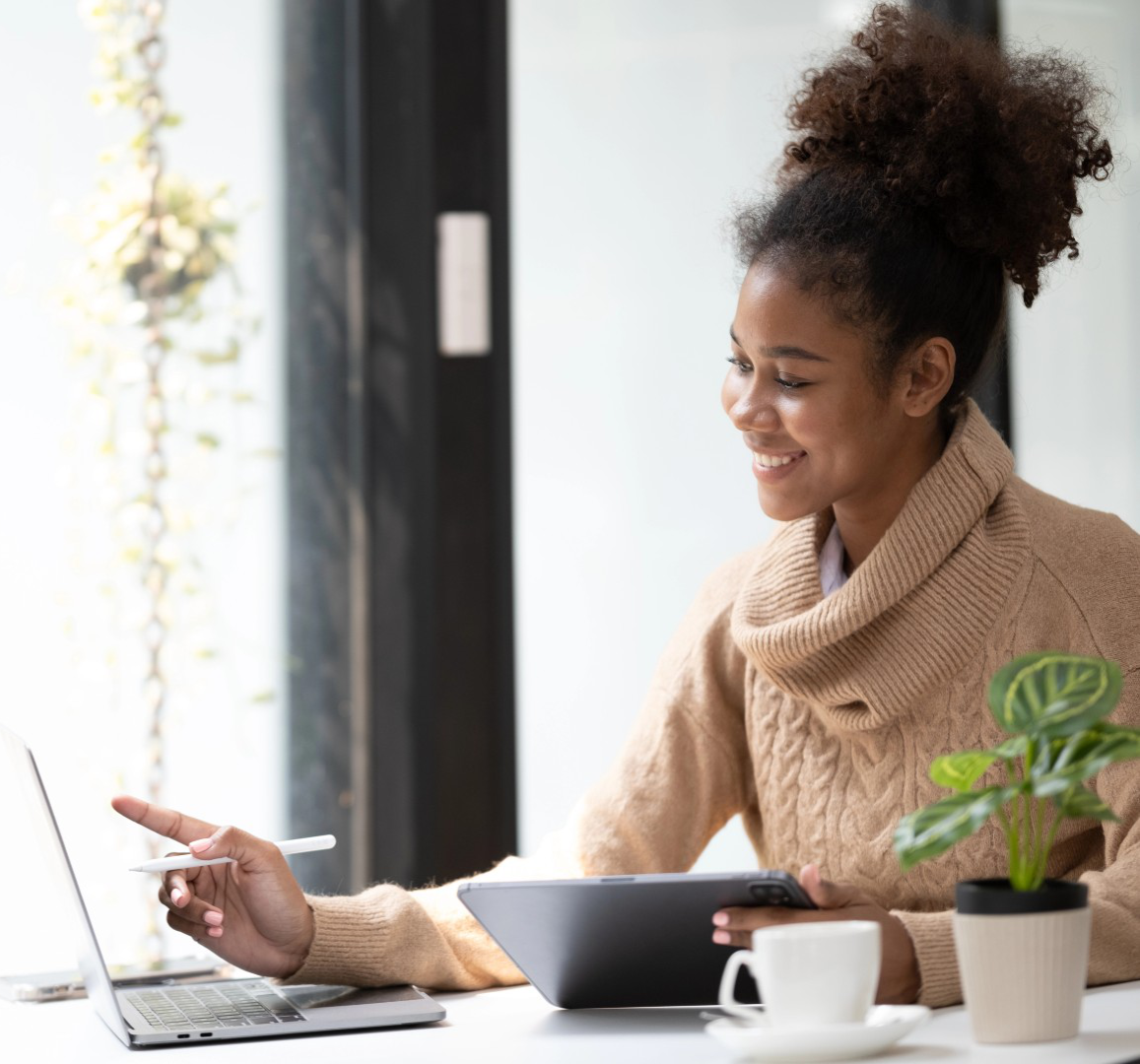 Smiling woman in a sweater pointing a stylus at a laptop while holding a tablet at a bright desk